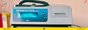 A grey Intoxilyzer 8000 breath alcohol testing device with a blue faceplate, displaying "Ready Mode Push Button to Start" on its green digital screen. It has a black mouthpiece tube coiled on top, a printer slot on the right, and buttons labeled "START TEST" and "POWER" with a green indicator light. To the left are various bottles and boxes. A yellow banner across the bottom contains the text "(505) 426-8711" and "GranoLaw.com".