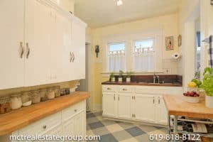 Vintage-inspired kitchen at 3758 31st Street in North Park featuring white cabinetry, warm wood butcher block countertops, blue and white checkered flooring, and double-hung windows with café curtains.