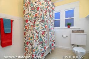 Craftsman bathroom at 3758 31st Street in North Park featuring a vintage clawfoot tub with a colorful paisley shower curtain, white wainscoting, yellow upper walls, and a double-hung window with privacy glass.