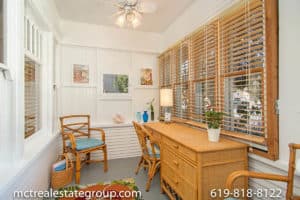 Bright and airy sunroom at 3758 31st Street in North Park utilized as a workspace, featuring white wood-paneled walls, a rattan desk and chair set, wood blinds, and a ceiling fan.