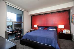 Modern bedroom at 321 10th Ave featuring a bold red accent wall, a dark platform bed with navy bedding, and a large window with views of the San Diego Central Library dome.
