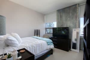 Modern bedroom at 321 10th Ave with a white bed, dark wood dresser with a TV, and a large window offering urban views of San Diego high-rises.