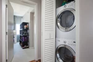 Interior hallway at 321 10th Ave featuring a stacked white front-loading washer and dryer in a closet with louvered doors, leading toward a carpeted room with a dark bookshelf.