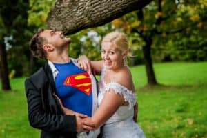 A romantic bride and groom dressed as superman are posing in front of a tree.