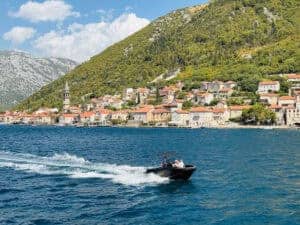 Panoramic view of a coastal town in Kotor Bay, Montenegro. Red-tiled roofs of stone houses rise along a hillside, with a bell tower visible in the foreground. The bay’s deep blue water is in the foreground, with a small black motorboat cutting through the water. Green hills and rocky mountains form the backdrop under a partly cloudy sky.