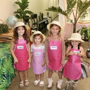 Four young girls in matching pink dresses and hats smiling indoors with plants.