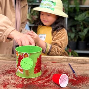 Child joyfully playing with red powder in a green bucket outdoors.