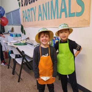 Two kids in aprons and hats at a party with animal theme.