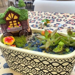 Miniature garden with succulent plants and decorative fairy house in a patterned pot.