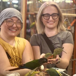 Two women smiling while feeding birds indoors.