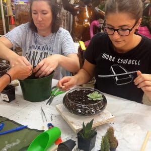 Two women craft succulent planters at a table filled with gardening tools and soil.
