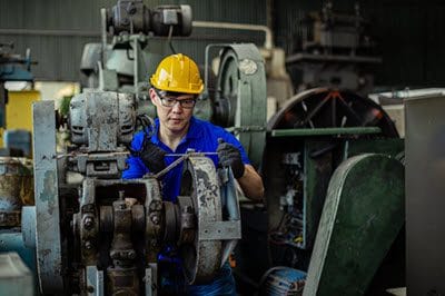Maintenance technician in a hard hat servicing industrial gear machinery, representing the hands-on cost of deferred maintenance