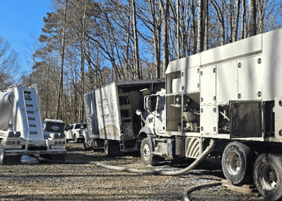 A large white CJGeo batch plant and support truck sit at a construction site in the woods.