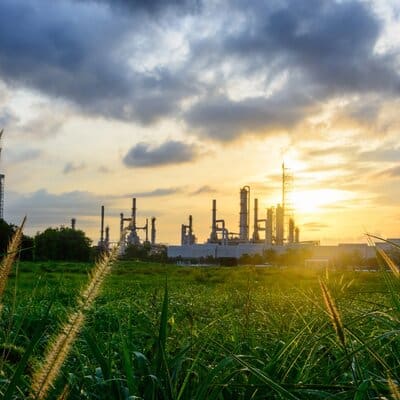 Industrial refinery or chemical plant silhouetted against a golden sunset sky beyond lush green grassland — illustrating the interface between industrial development and natural habitat requiring air quality permitting, environmental impact assessment, and regulatory compliance consulting by EDGE Engineering.