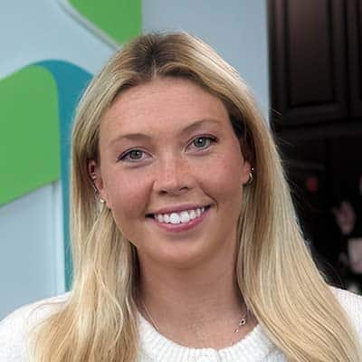 A young woman with long blonde hair smiles at the camera. She is wearing a white top, standing indoors with a blue and green abstract background and dark cabinets behind her.