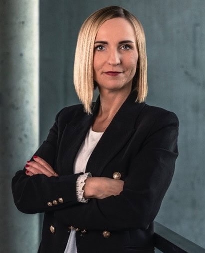 Zdjęcie przedstawia pewną siebie kobietę w czarnym garniturze, stojącą z założonymi rękami, na tle nowoczesnej ściany, promującą wydarzenie PHD RISK Summerschool i tematykę bezpieczeństwa i ryzyka.