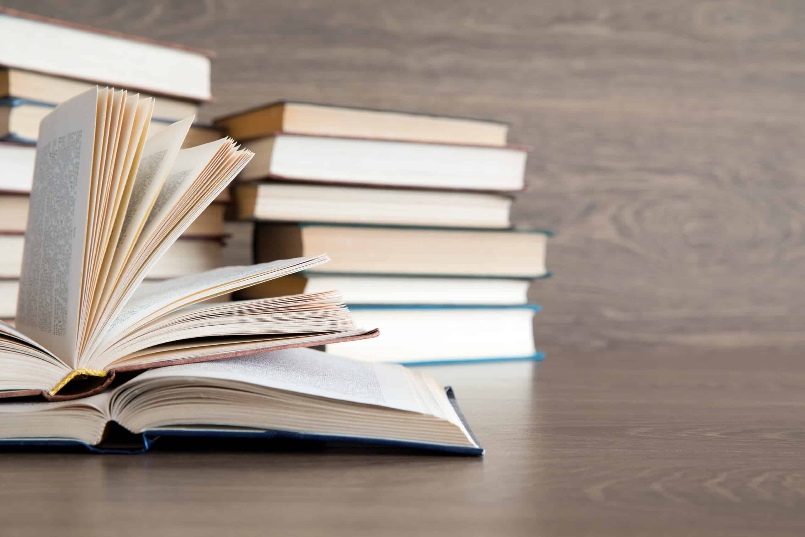 A book open with pages spread in foreground with a pile of books in background