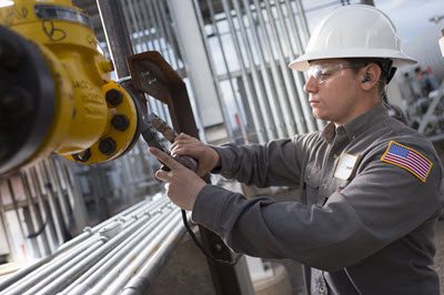 Maintenance technician in a hard hat and safety glasses uses a large wrench to adjust an industrial valve on piping inside a plant.