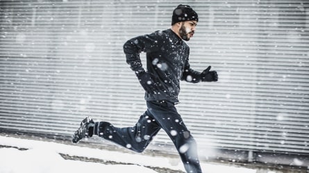 Athlete in black running gear running on a sidewalk in heavy snow.