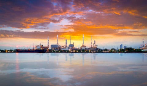 Petrochemical refinery complex illuminated at sunset with vivid orange and purple sky reflected in calm waterway — industrial facility subject to air quality permitting, emissions monitoring, and Clean Air Act compliance consulting by EDGE Engineering.