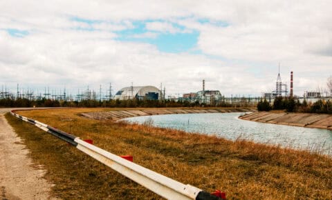 Chernobyl Nuclear Power Plant with its New Safe Confinement arch structure visible across an industrial canal, surrounded by flat terrain and power transmission infrastructure — site of the 1986 nuclear disaster used to illustrate industrial facility environmental risk and remediation challenges.