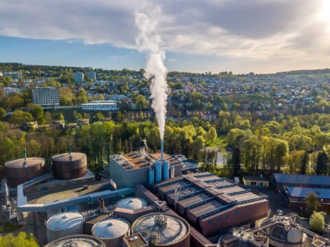 Aerial view of a wastewater treatment or industrial processing facility with digesters, storage tanks, and an active steam emissions stack adjacent to a reside…Aerial view of a wastewater treatment or industrial processing facility with digesters, storage tanks, and an active steam emissions stack adjacent to a residential community and green tree buffer — urban industrial facility subject to air quality permitting, odor control compliance, and environmental regulatory consulting by EDGE Engineering.