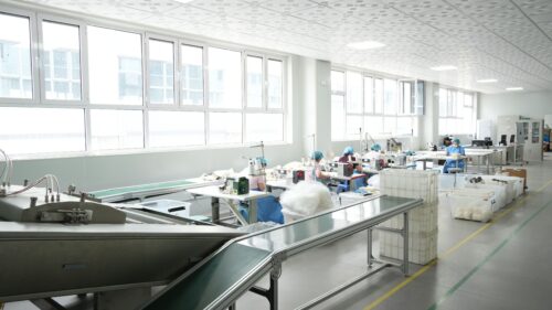 Workers wearing protective headwear operate assembly workstations along a conveyor belt in a bright manufacturing facility.