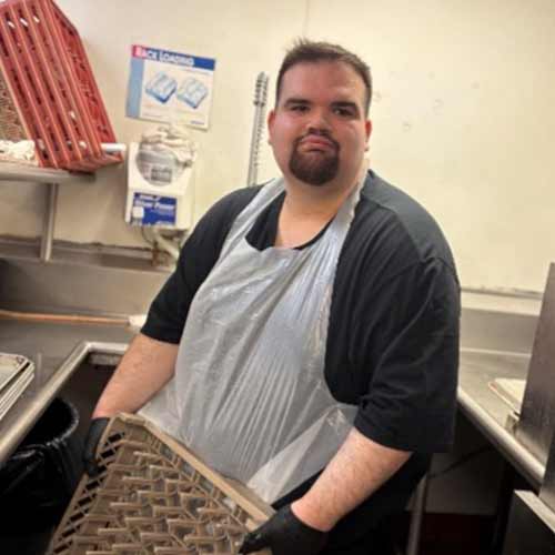 A man wearing a plastic apron and black gloves stands in a commercial kitchen, holding a dish rack near a sink. There are kitchen supplies and a sanitation sign visible in the background.