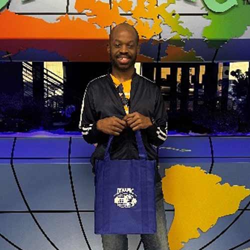 A person smiles while standing indoors, holding a blue tote bag that reads "TITANIC FAN CLUB." A colorful map of the world is displayed on the wall behind them.