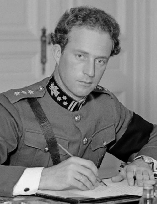 Koning Leopold III zit achter zijn bureau in het paleis te Laken, gekleed in uniform, met documenten voor zich.