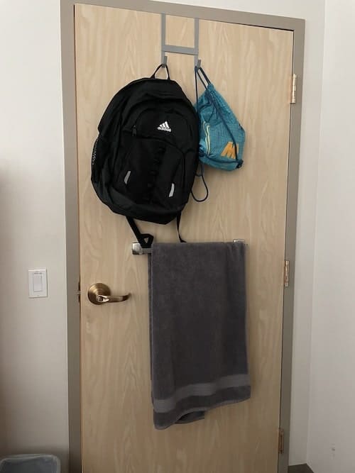 A dorm room door with over-the-door hooks for backpack and shower items, plus a towel bar