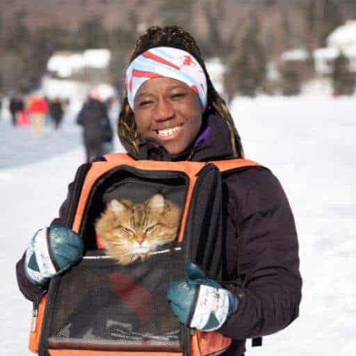 african american woman in the snow wearing a red white and blue headband with skier pattern and black jacket carrying a brown tabby cat in an orange backpack