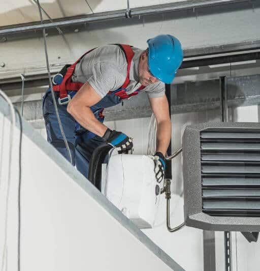 man in work overalls gray tshirt and blue hard hat fixing a heater.
