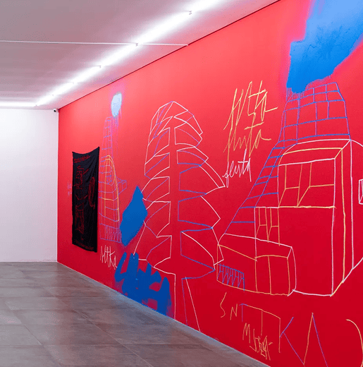 Uma parede de galeria vermelha vibrante exibe desenhos coloridos de estruturas e árvores, um tecido preto e texto como "MUTA MUTA SEUTA", sob luzes.