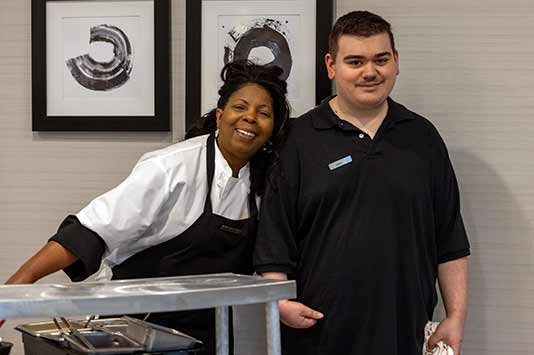 Two hotel staff members, one wearing a chef coat and apron and the other in a black polo, stand smiling together near a metal food cart. Abstract black-and-white art hangs on the wall behind them.