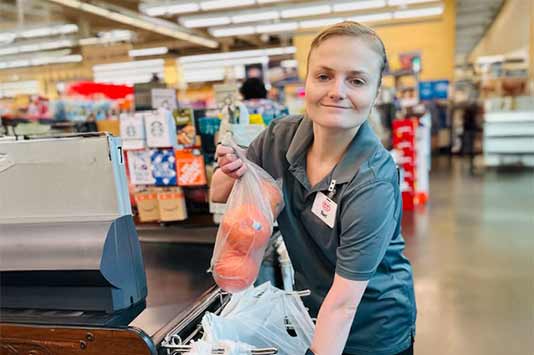 A grocery store employee smiles while bagging groceries at a checkout counter, holding up a plastic bag with produce inside. Shelves and products are visible in the background.