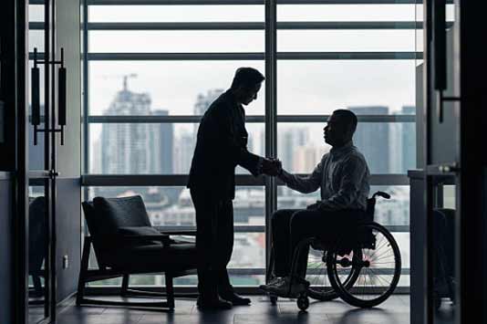 Two men greet each other in an office with large windows; one is standing and the other is seated in a wheelchair. The city skyline is visible in the background.