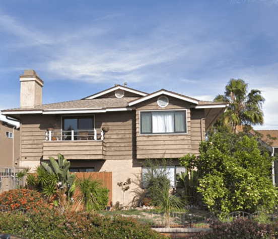Two-story brown wood-sided home at 3689 Texas Street in San Diego featuring a small upper balcony and lush landscaping with tropical plants and a lemon tree.