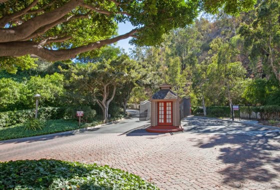 Gated community entrance at 5474 Caminito Borde featuring a brick-paved driveway, a security kiosk with red doors, and lush green hillside landscaping.