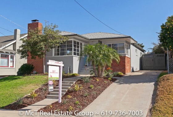 Exterior view of a contemporary grey home at 1627 Granada Avenue in South Park featuring a prominent red brick chimney, wrap-around corner windows, a tiered garden bed with succulents, and a paved driveway under a clear sky.