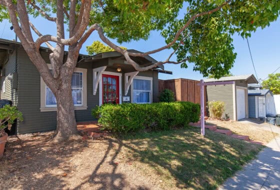 Exterior view of a home with olive green siding, a red front door, a large shade tree in the front yard, and a real estate sign post near the driveway at 2221 Wightman Street in North Park.