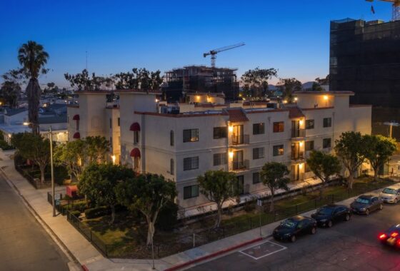 Exterior view of a multi-story stucco apartment building at dusk with lit windows, red awnings, surrounding trees, and cars parked along the street at 205 Ohio St Unit 304 in North Park.