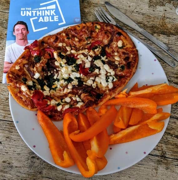 Goats cheese pizza with raw orange pepper sticks on a white spotty plate on a wooden table with cutlery and do the unthinkable booklet