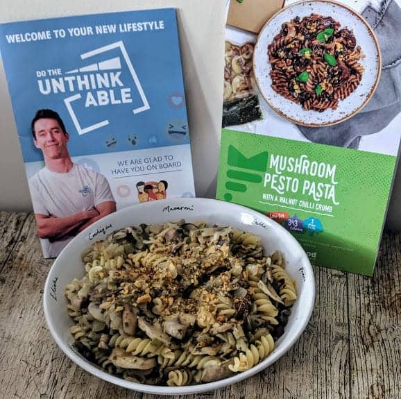 Mushroom pesto pasta with a walnut chilli crumb in a bowl on a wooden table with booklet and packaging.