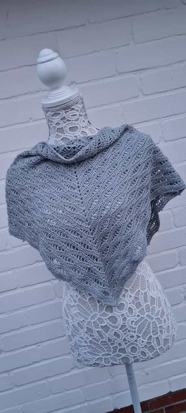 Handgearbeiteter Grau gestrickter Poncho auf stilvollem Dekorationsständer vor Ziegelwand.