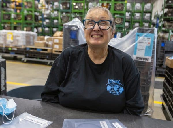 An older woman wearing glasses and a black shirt with a "Dynamic" logo smiles broadly in a warehouse setting. There's a table in front of her with packages and a face mask. Rows of shelves and bins are visible in the background.