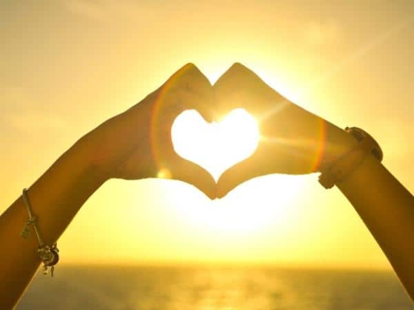 a heart made with a ladies hands at sunset with sun shining through the heart