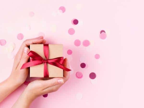 Two hands holding gift box above pink surface with pink confetti