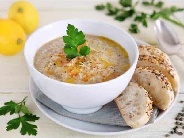 A white bowl of aubergine soup with bread on the side of the plate