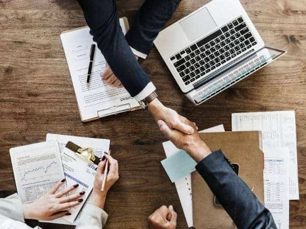 Three people at a meeting table with documents and a laptop. Two are shaking hands across the table.
